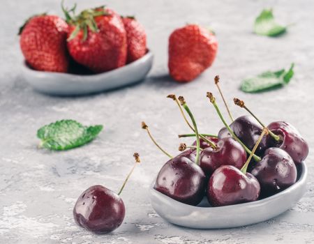 Fresh ripe cherries in small trendy plate. Cherry in gray plate, strawberry on background, mint leaves on gray concrete background