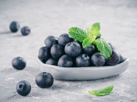 Fresh ripe blueberries in small trendy plate. Blueberry in gray plate and mint leaves on gray concrete background. Copy space.