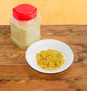 White dish with rice cooked with carrots and spices against of the transparent plastic container with uncooked rice on the surface of the old wooden planks
