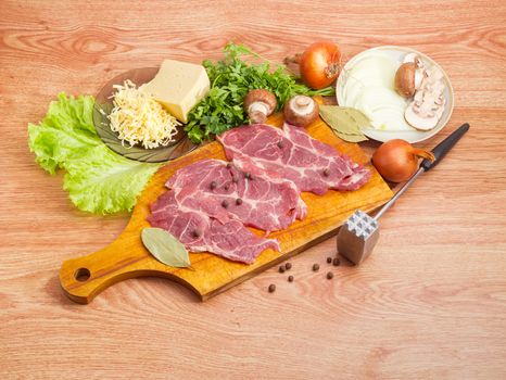 Sliced uncooked pork neck on the wooden cutting board, cheese, mushrooms, onion, greens and spices for cooking french-style meat, beside meat tenderizer on a wooden surface
