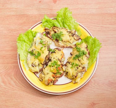 Top view of several pork chops baked with onion, mushrooms and cheese and decorated with twigs of the parsley and lettuce leaves on the yellow and white dish on a wooden surface
