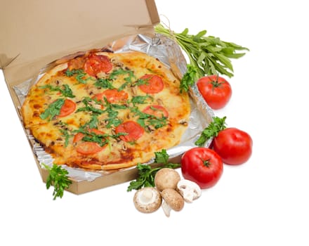Cooked round pizza with tomatoes, mushrooms and arugula wrapped in aluminum foil in the open cardboard box and fresh tomatoes, mushrooms, parsley and arugula beside on a light background
