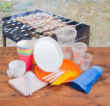 Different disposable plastic and paper cutlery, paper napkins and roll of disposable garbage bags on the old wooden planks against the background of barbecue grill with grilled skewered meat outdoors
