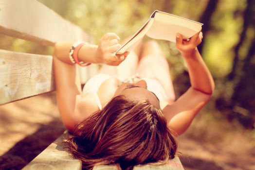 Young woman reading a book while relaxing in the forest. She is laying on bench and enjoying.