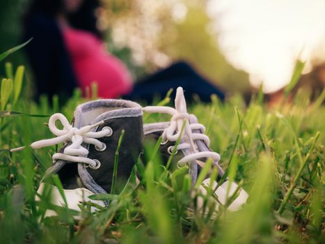 Close up view of little children shoes in grass and blurred silhouette of pregnant woman on background. Shallow DOF, focus on foreground. Pregnancy concept. Copy space.