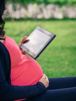 Close up view of pregnant belly and tablet or e-reader. Pregnancy relaxing outdoors concept. Copy space. Vertical.