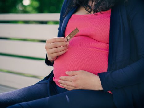 Pregnant woman sitting outdoors and hold piece of chocolate near stomach