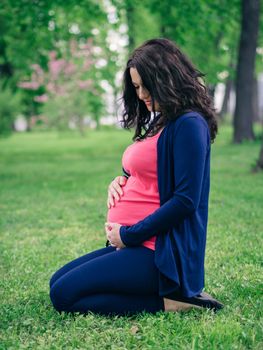 Pregnant woman siting on grass in park. Looks at belly. Hand on abdomen. Vertical. Copy space.