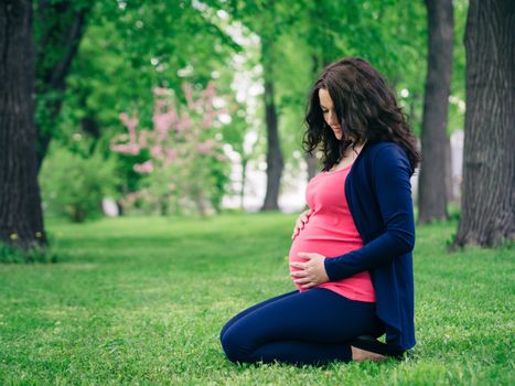 Pregnant woman siting on grass in park. Looks at belly. Hand on abdomen. Copy space.