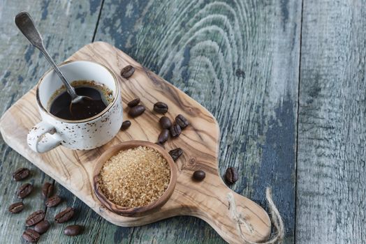 Black coffee in old coffee cup with a silver teaspoon, spilled coffee beans and cane sugar in a wooden bowl on the old wooden table