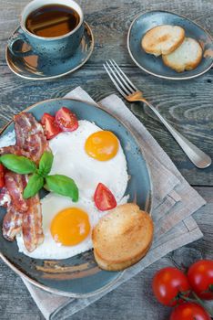 Breakfast consists of fried eggs,  bacon, tomato, toast and a cup of coffee on the old wooden table