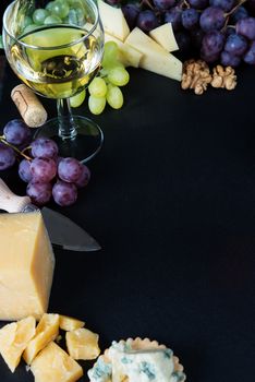 White wine, various types of cheese, walnuts and grapes on a black slate stone background