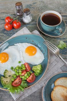 Breakfast consists of fried eggs,  bacon and various vegetables (tomato, green pea, broccoli, lettuce), toast and a cup of coffee on the old wooden table; vertical image