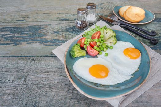 Breakfast consists of fried eggs,  bacon, various vegetables (tomato, green pea, broccoli, lettuce) and toast on the old wooden table; with copy-space