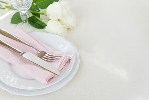 Beautiful decorated table with white plates, crystal glass, linen pink napkin, cutlery and white rose flowers on tablecloths, with space for text