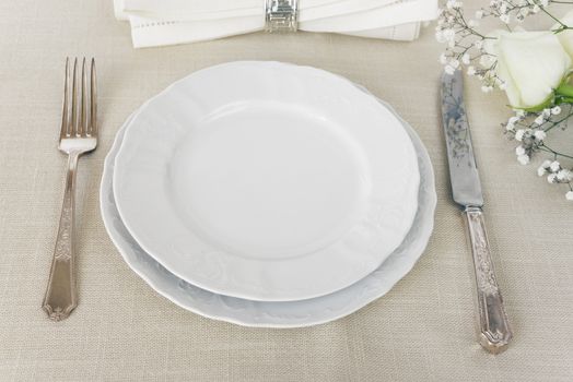 Beautiful decorated table with empty white plates, linen napkin, cutlery and white rose flowers on tablecloths, with space for text