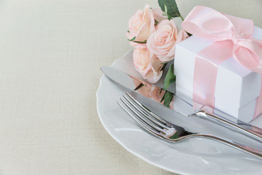 Beautiful decorated table with white plates, gift box with a pink ribbon, cutlery and pink rose flowers on tablecloths, with space for text