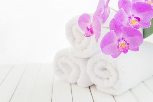 SPA concept: stack of three rolls of white fluffy bath towels with violet orchid flower on the background of white boards