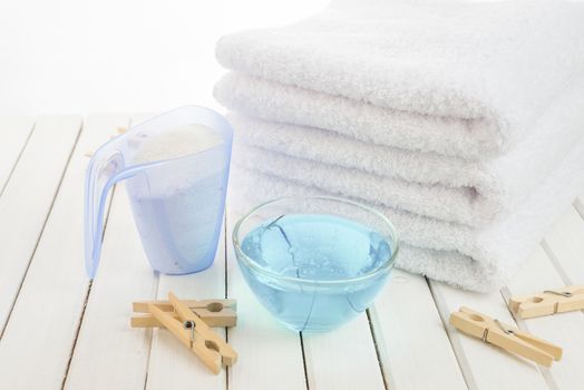 Stack of three white fluffy bath towels, washing powder in measuring cup, blue fabric softener in a transparent glass bowl and wooden clothespins on the background of white boards