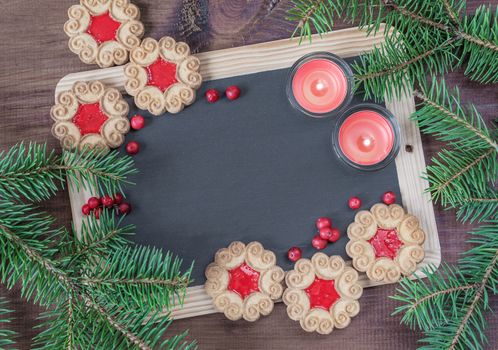 Blank chalkboard surrounded by green fir branches, two red burning candles and few cookies with berry jam, top view, flat lay