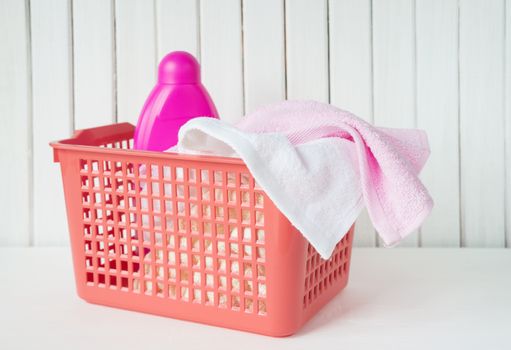 White and pink towels and detergent in pink bottle are in a red plastic laundry basket on the white wooden background