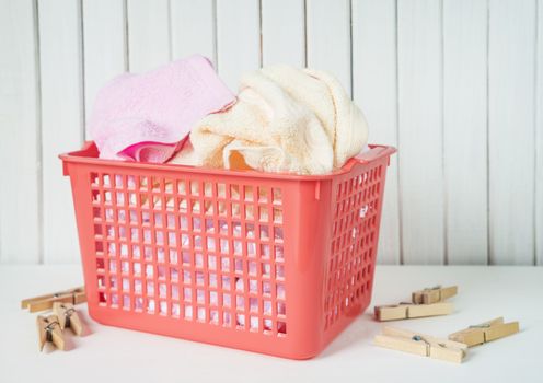 White, beige and pink towels in a red plastic laundry basket and wooden clothespins are on the white wooden background