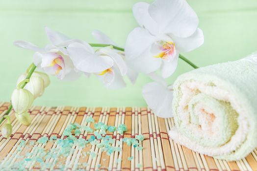 Spa concept: bath towels and bath salt on a natural mat  and a branch of white orchid on a green background