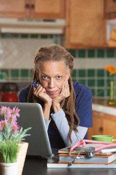 Thoughtful medical student studying on laptop with books and stethoscope in her kitchen