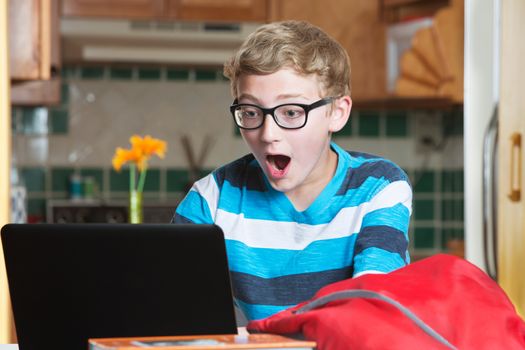 Surprised boy in eyeglasses and striped shirt using laptop with red backpack