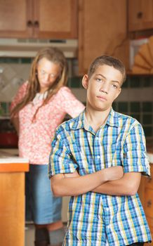 Teenaged son stands with arms crossed in the kitchen after having disagreement with him mother