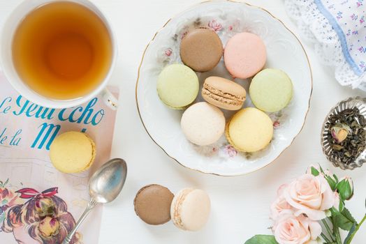 Multicoloured macaroons on beautiful porcelain plate, cap of tea and rose flowers on white background; top view, flat lay