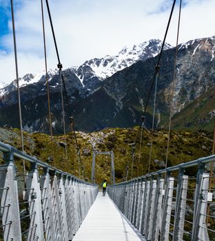 Hiker walking across suspension bridge with bright green backpack. Jagged snow capped mountains in the background.
