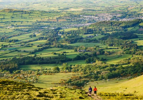 Two women walking away off of hill top in sports gear. Farmland and town in the distance. Taken in Wales during golden hour.
