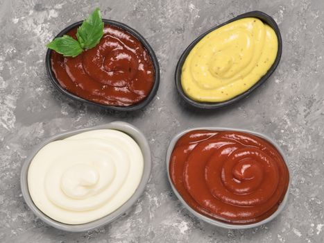 Top view of classic sauces set in trendy plates on gray concrete background. Sauces set - salsa, mustard, ketchup, mayonnaise, and basil for dressing. Flat lay.