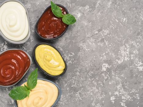 Top view of classic sauces set in trendy plates on gray concrete background. Sauces set - salsa, mustard, ketchup, mayonnaise, cheese sauce, and basil for serve. Flat lay. Copy space.