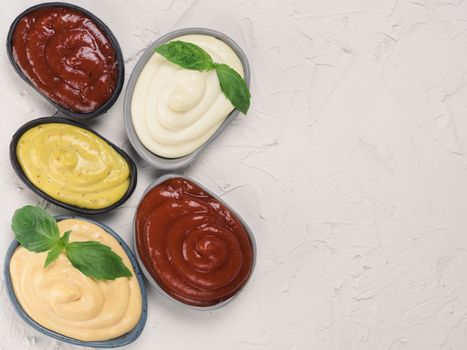Top view of classic sauces set in trendy plates on white concrete background. Sauces set - salsa, mustard, ketchup, mayonnaise, cheese sauce, and basil for serve. Flat lay. Copy space.