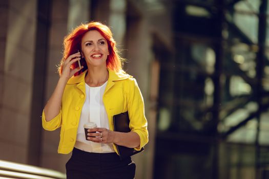 Smiling successful businesswoman using smartphone on a coffee break in office district. Looking away.