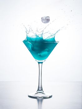 Blue cocktail in martini glass with ice cube splashing into liquid against white background. Blue curacao cocktail with splash on white.