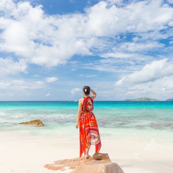 Traditinaly dressed local woman wearing long floral summer dress and hat looking at sea on Anse Patates beach, La Digue Island, Seychelles. Summer vacations on picture perfect tropical island concept.