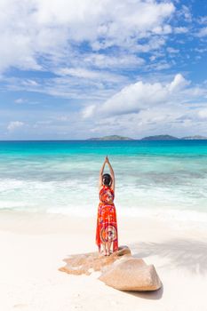 Traditinaly dressed asian tourist woman wearing long red floral summer dress doing yoga by sea on Anse Patates beach, La Digue Island, Seychelles. Summer vacations on picture perfect tropical island.