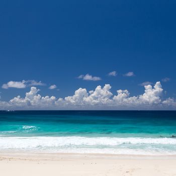 Empty tropical beach on Seychelles featuring levels of white sand, emerald water, dark blue water, and dark blue sky with white clouds. Copy space on the sky.