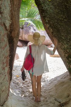 Rear view of barefooted woman wearing white loose tunic over bikini and beach hat on Anse Lazio beach on Praslin Island, Seychelles. Summer vacations on picture perfect tropical beach concept.