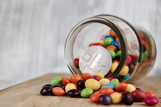 Digital currency physical silver Litecoin coin in jar near colorful sweets on the wooden board.