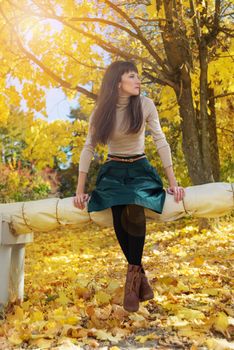 Slim brunette girl with long hair in a green skirt and beige turtleneck sitting in autumn park and looking away