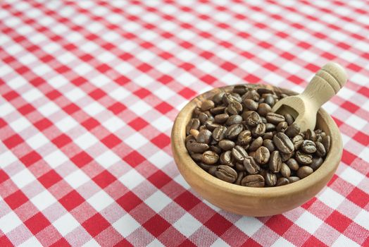 Coffee beans in the wooden bowl and wooden scoop are on a background of red and white checkered cloth