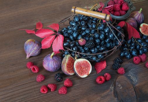 Black grapes in a basket and ripe figs, red raspberries and blackberries with autumn leaves on the wooden background