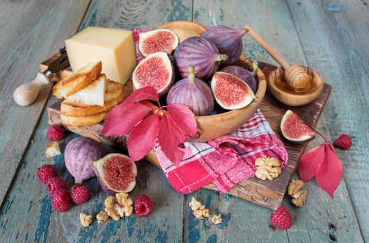 Ripe figs in a wooden bowl, red raspberry, cane sugar, honey and a checkered napkin on old cutting board as well as autumn leaves lie on the old wooden table