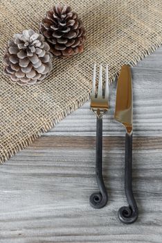 Steel fork and knife handmade and two pine cones are located on the rough wooden table, covered with coarse matting