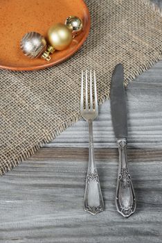 Steel fork and knife and Christmas balls on a ceramic plate are located on the rough wooden table, covered with coarse matting