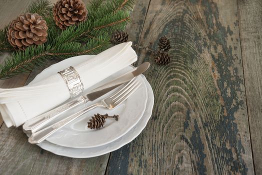 Christmas table: silver knife, fork and linen napkin lie on the white dinner plate, as well as green spruce branch and cones which is located on a old wooden table, with space for text